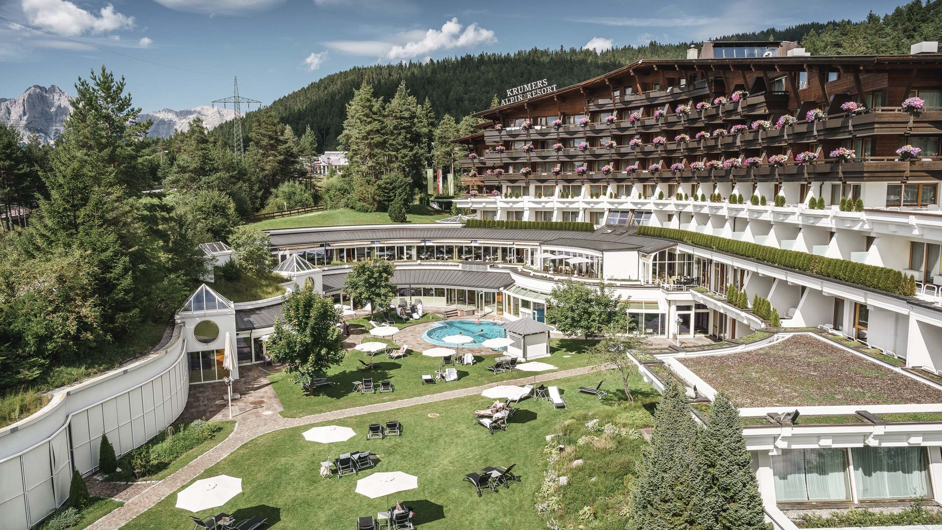 Ihr Hotel in Seefeld: Krumers Alpin Alpenresort mit Garten, Pool und Bergen im Hintergrund bei klarem Wetter